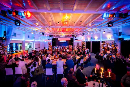 Gezellige feestzaal gevuld met mensen aan tafels, verlicht door sfeervolle kleurrijke verlichting. De zaal heeft een open houten plafond en is versierd met bloemen en kaarsen, wat zorgt voor een warme en uitnodigende ambiance.